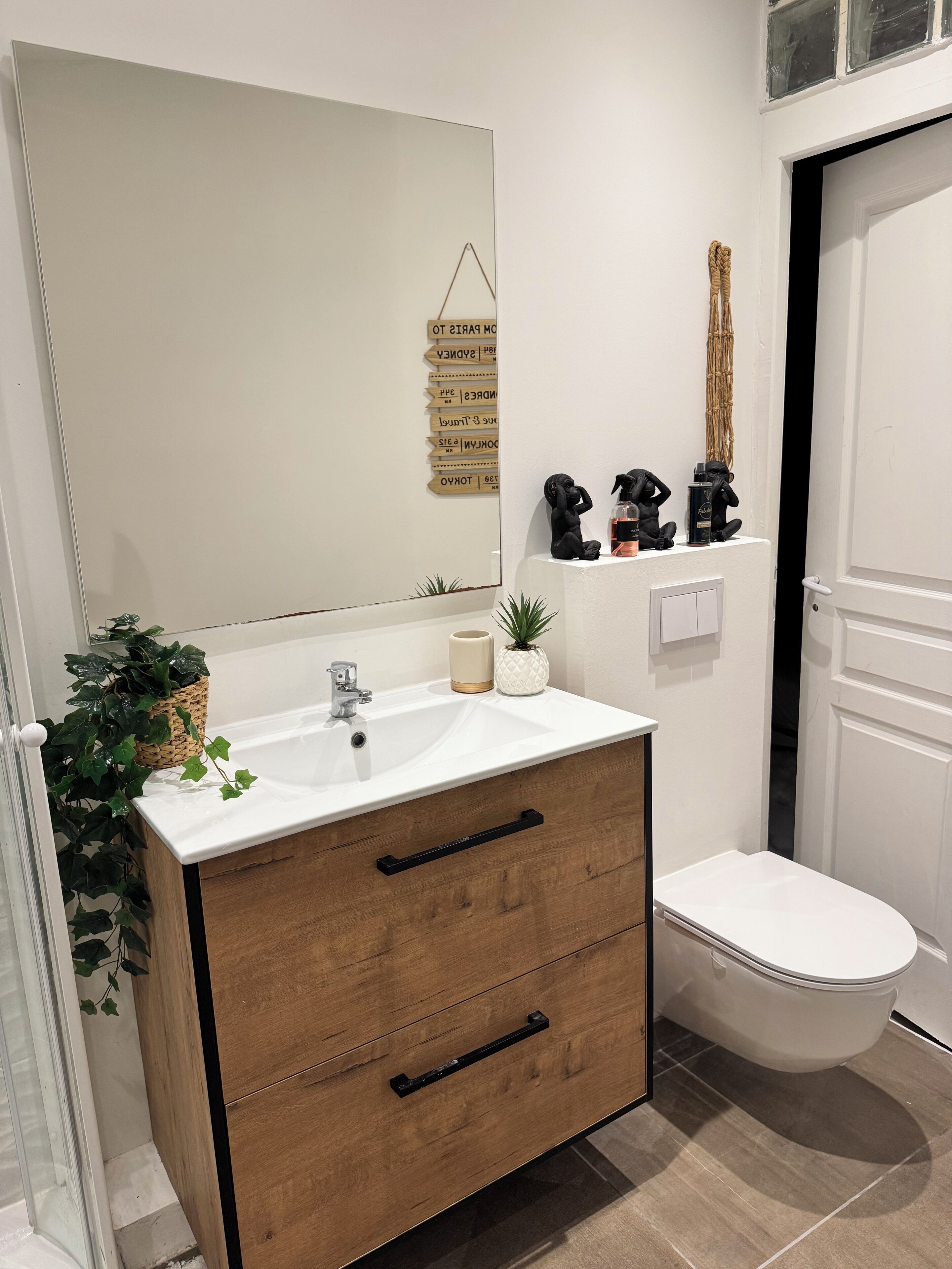 Une salle de bain moderne avec un meuble-lavabo en bois, un grand miroir, des toilettes et une petite étagère ornée d’objets décoratifs.