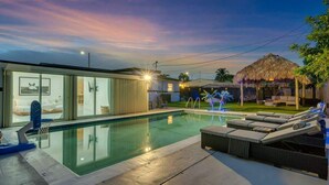 Uma área de piscina moderna com um espaço envidraçado, espreguiçadeiras e uma estrutura com telhado de palha.