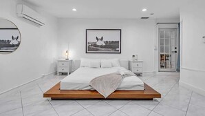 Quarto moderno com cama de plataforma em madeira, roupa de cama branca, espelho redondo, fotografia emoldurada e mesa de cabeceira com abajur.
