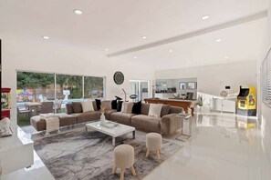 A modern living room with a sofa, ottomans, and a coffee table. There is a kitchen area with a dining table and chairs visible through an open door. - Lux Miami Villa Heated Pool 10 min to Beach (North Miami Beach)