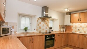 A modern kitchen with wooden cabinets, a built-in oven, a microwave, and a window with blinds. - Hay Loft (Anstruther)