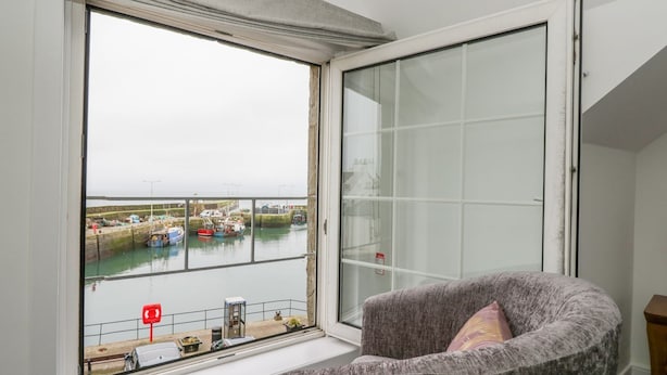 Ein Zimmer mit großem Fenster mit Blick auf eine Marina, ein grauer Sessel mit Kissen und eine Schiebetür.