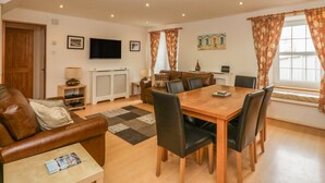 A cozy living room with a brown leather sofa, a wooden dining table with chairs, a TV mounted on the wall, and a window with curtains. - Flour Loft (Anstruther)