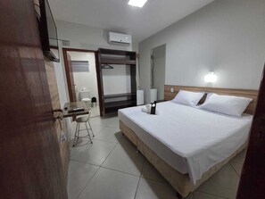 A hotel room with a bed, a desk with a chair, a wardrobe, a toilet, and an air conditioning unit.