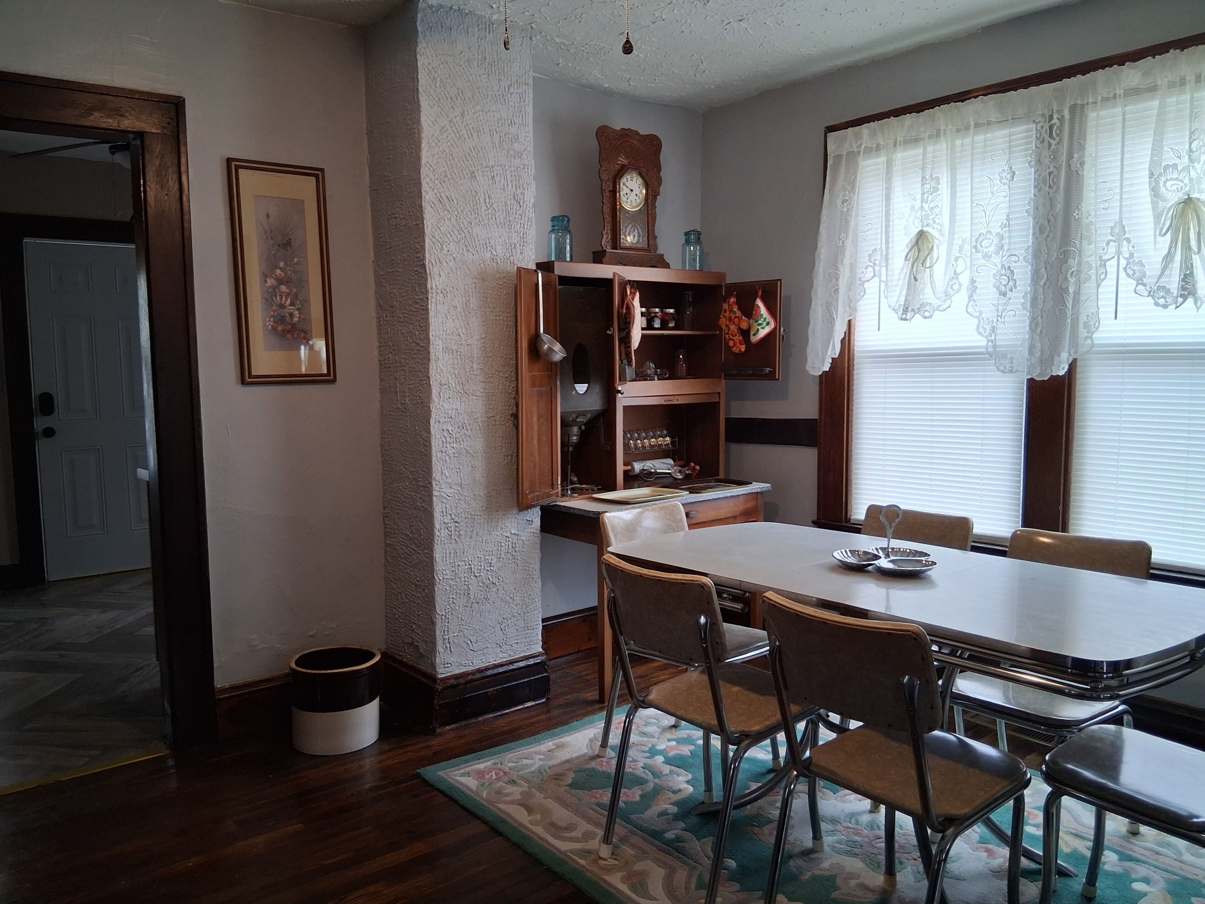 A dining room with a wooden table, chairs, a patterned rug, a clock, and a window with curtains.