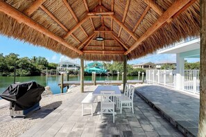 A covered patio with a thatched roof, a grill, and a view of a lake and buildings. - Dockside Paradise | Brand New, Large Dock, Private Pool, Tiki Hut, Walk to Sombrero Beach (Marathon)