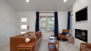 A modern living room with a brown leather sofa, a wooden coffee table, a small side table, a blue armchair, and a fireplace. - Four Cabins near each other- King Beds, AC, Firepit, SMores, Sledding (Angel Fire)