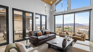 A spacious living room with a leather sofa, a coffee table, and a view of a mountainous landscape through large windows. - 2 Min to Jordanelle Gondola Hot Tub Spa Suite Movie Room & Mountain Views Skyridge Summit by Avantstay (Heber City)