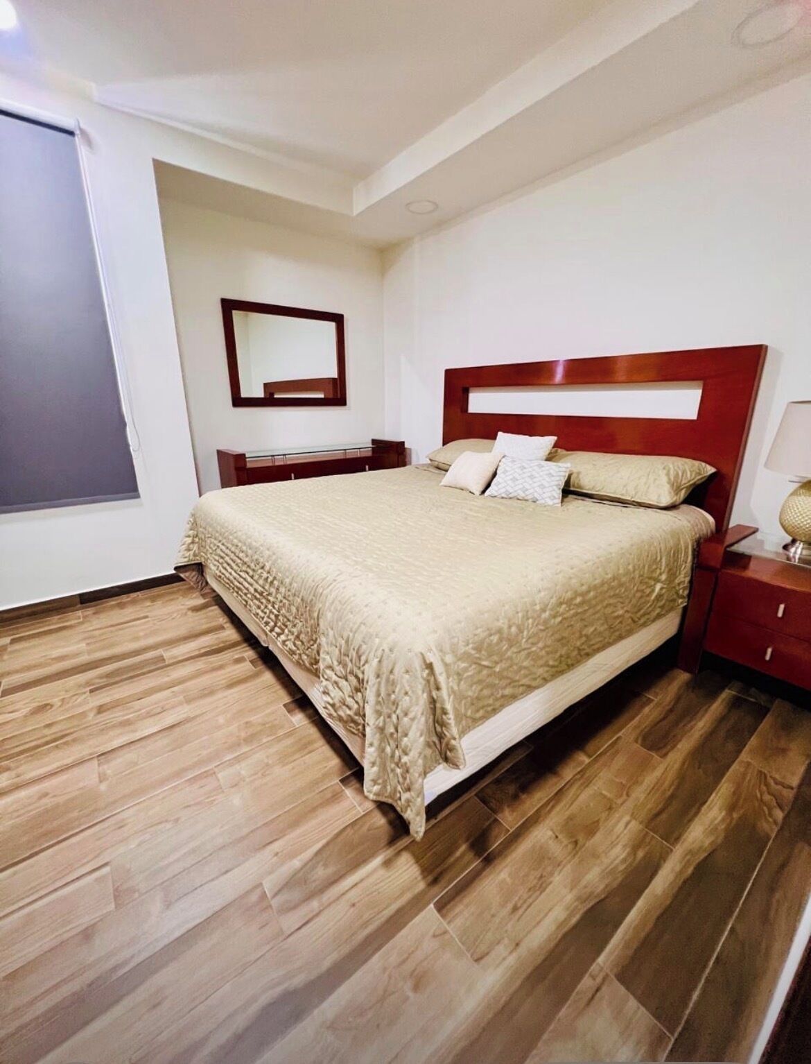 A bedroom with a large bed, wooden flooring, a nightstand, a bedside table, and a mirror on the wall.