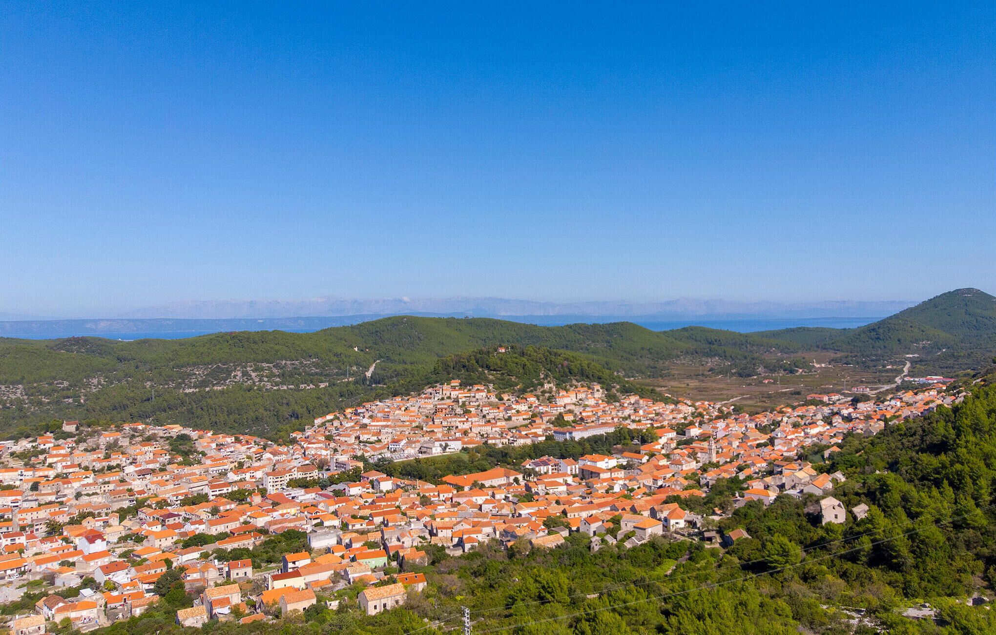 A coastal town with densely packed buildings, a clear blue sky, and a hilly backdrop.