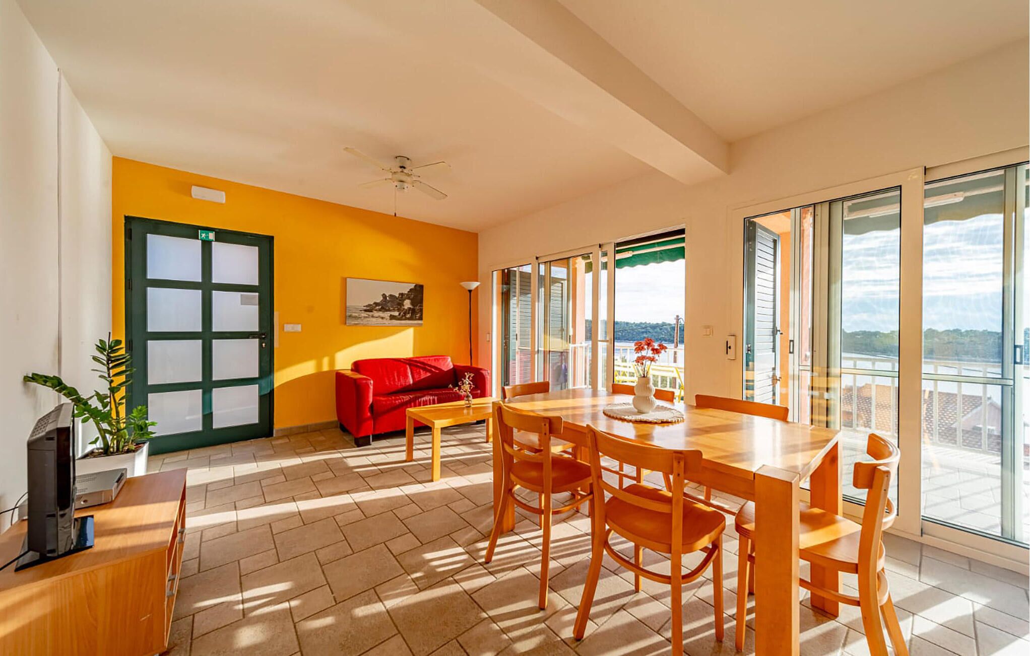 A bright living room with a red sofa, a wooden dining table with chairs, a ceiling fan, and a large window offering a view of the sea.