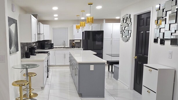 Una cocina moderna con armarios blancos, una isla central y un refrigerador negro. Hay tres taburetes dorados y una mesa con cubierta de vidrio.