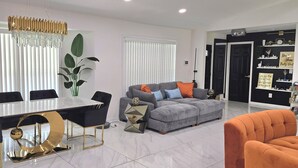 Una sala de estar moderna con comedor, que incluye una mesa de comedor blanca, sillas negras, un sofá gris con cojines anaranjados y una pared blanco y negro con estanterías.