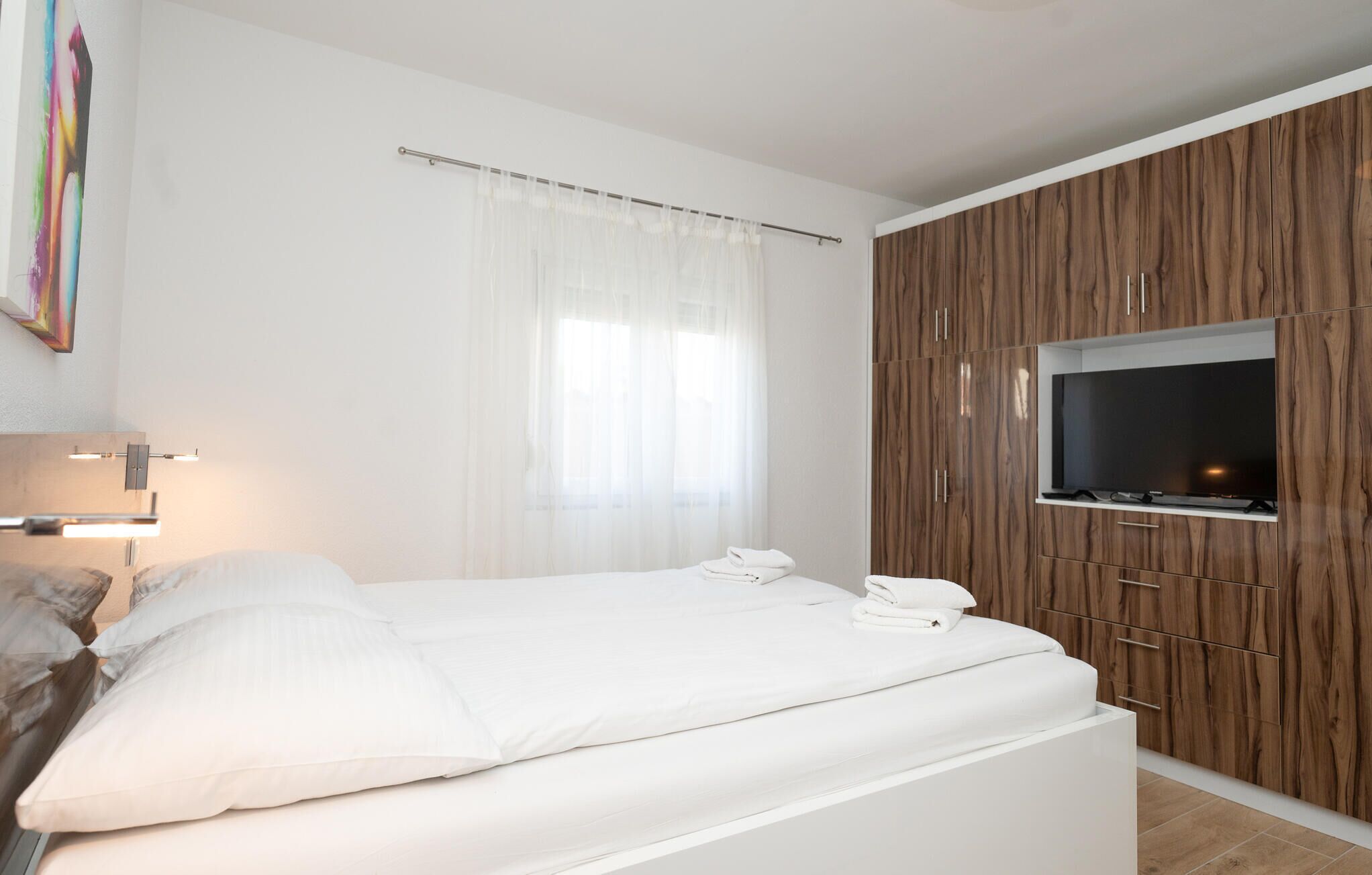 Una camera da letto moderna con un letto, una TV montata su un mobile in legno e una finestra con tende leggere.