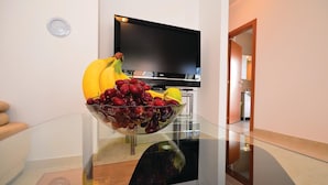 A modern living room with a glass coffee table holding a fruit bowl, a flat-screen TV, and a wooden door leading to another room.