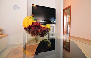Un salon moderne avec une table basse en verre sur laquelle repose un panier de fruits, un téléviseur à écran plat et une porte en bois donnant sur une autre pièce.