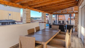 An outdoor patio with a wooden table and wicker chairs, a pool in the background, and a view of buildings and hills. - Gorgeous home in Posedarje (Posedarje)