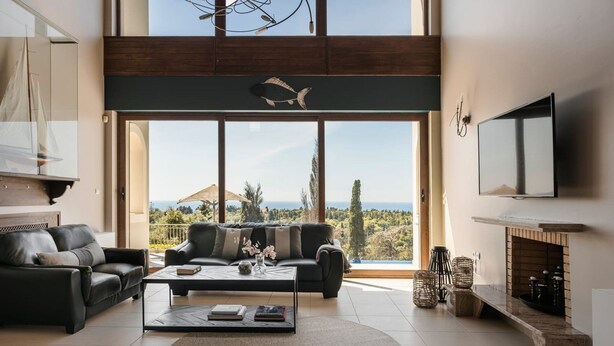 A spacious living room with a large window overlooking a scenic view, a fireplace, and a flat-screen TV mounted on the wall. - Villa Phoebeia, Wonderful Sea Views, Outdoor Infinity Pool, Antipata, Kefalonia (Kefalonia)