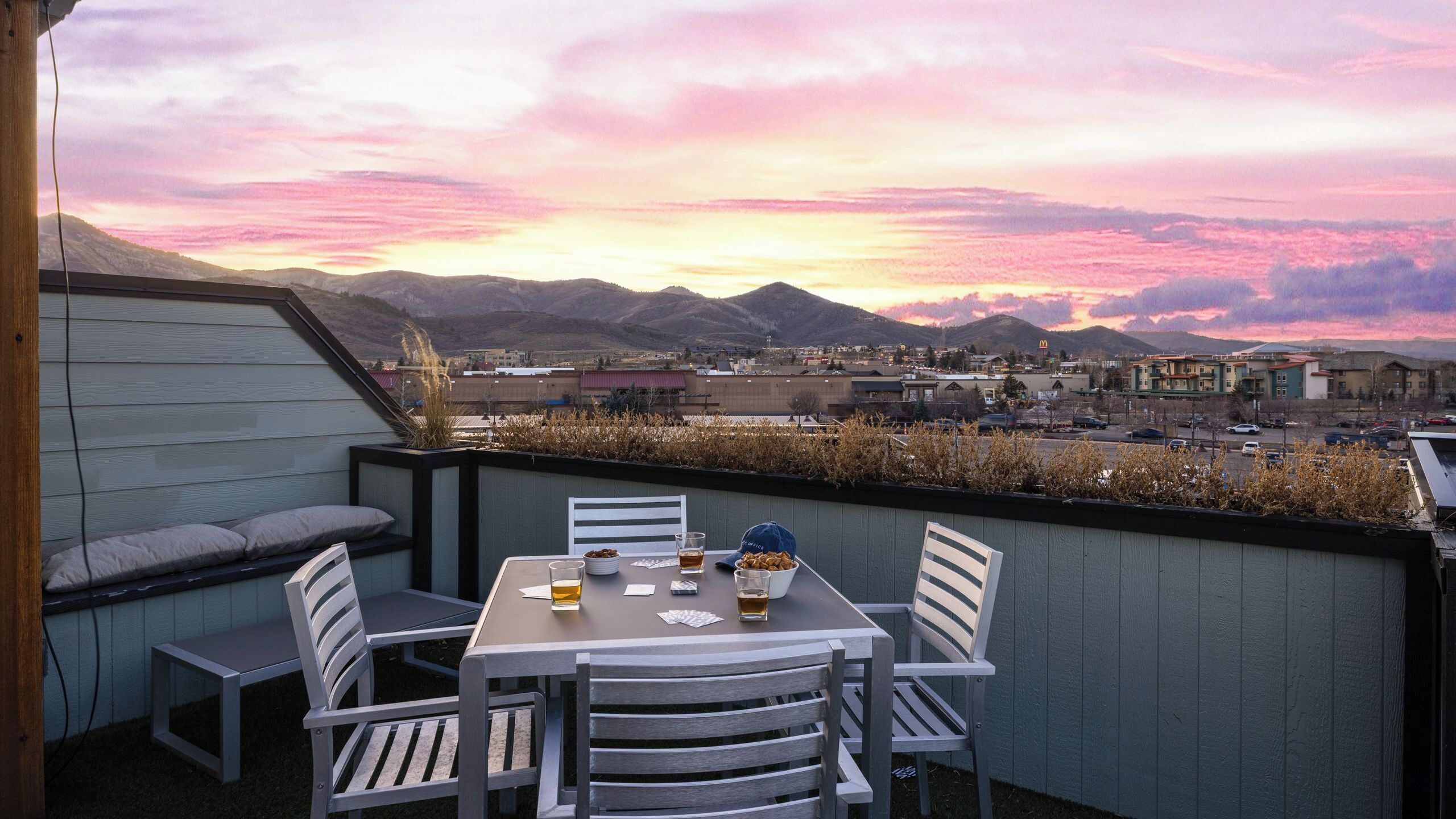 A rooftop patio with a table set for two, surrounded by chairs, overlooking a cityscape with mountains in the distance.