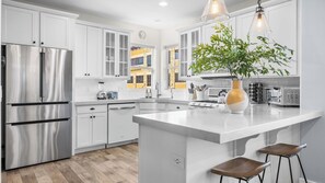 Cozinha moderna com armários brancos, eletrodomésticos de aço inoxidável, uma ilha central, banquetas e um vaso com planta.
