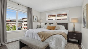 A bedroom with a large bed, a bench, a nightstand, a window with curtains, and a view of buildings outside. - Rooftop Deck w/ Hot Tub Near Dining & Ski Slopes The Lookout by Avantstay (Park City)