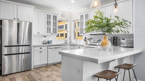 A modern kitchen with white cabinetry, stainless steel appliances, a central island with stools, and a view of a yellow building through the windows. - Rooftop Deck w/ Hot Tub Near Dining & Ski Slopes The Lookout by Avantstay (Park City)