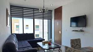 A modern living room with a sectional sofa, a glass coffee table, a television mounted on the wall, and a view of buildings outside. - Rio Nilo Apartments (Mazatlán)