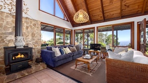 A cozy living room with a stone fireplace, a blue sofa, wicker furniture, and large windows offering a view of the outdoors.
