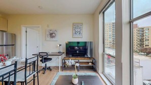 A modern living room with a large window, a flat-screen TV, a dining area with a table and chairs, and a small potted plant. - Stylish Home With Free ParkingFA (Stamford)