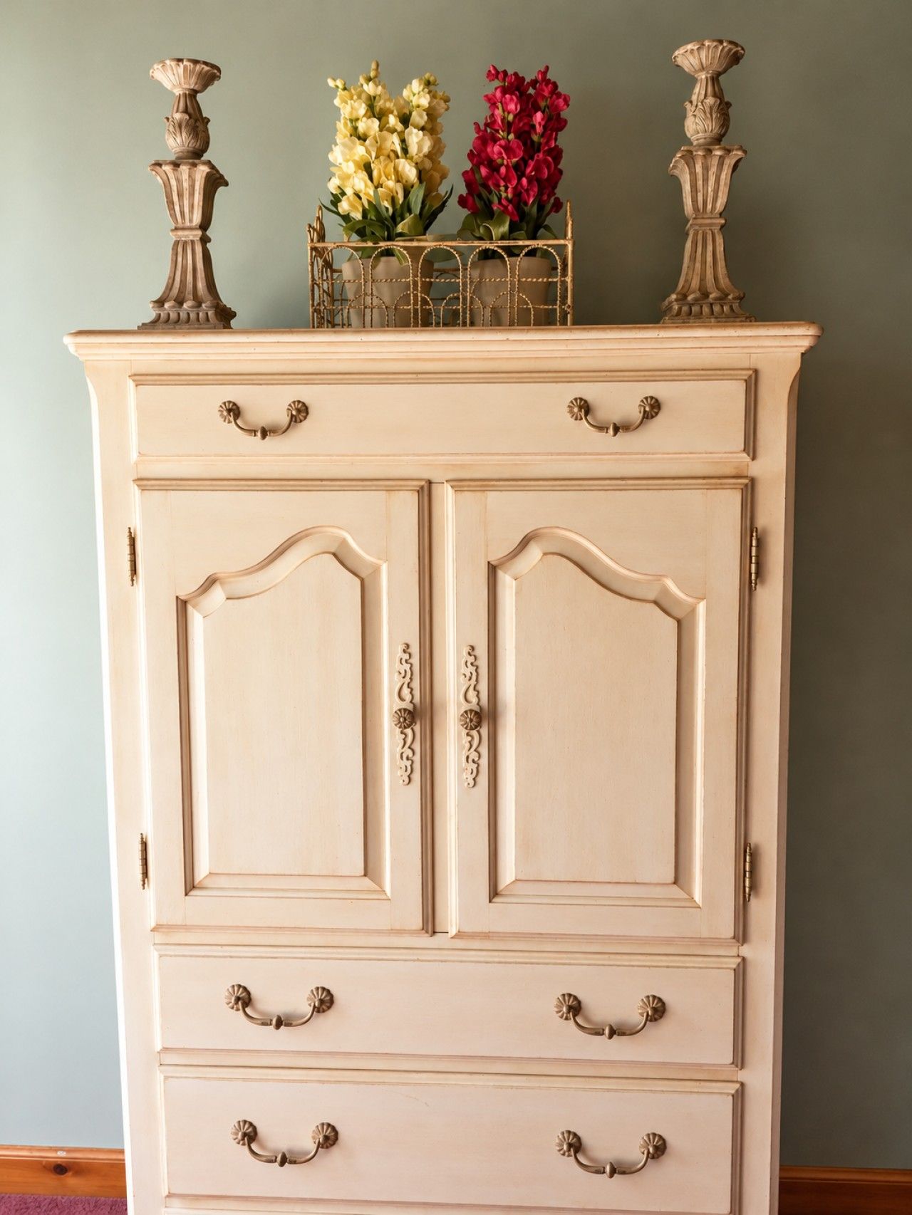 Um armário cor de creme com duas portas e várias gavetas, coroado por um suporte de metal decorativo com flores.