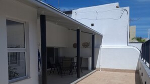 Uma área externa coberta com mesa e cadeiras, uma janela e um edifício branco com telhado plano.