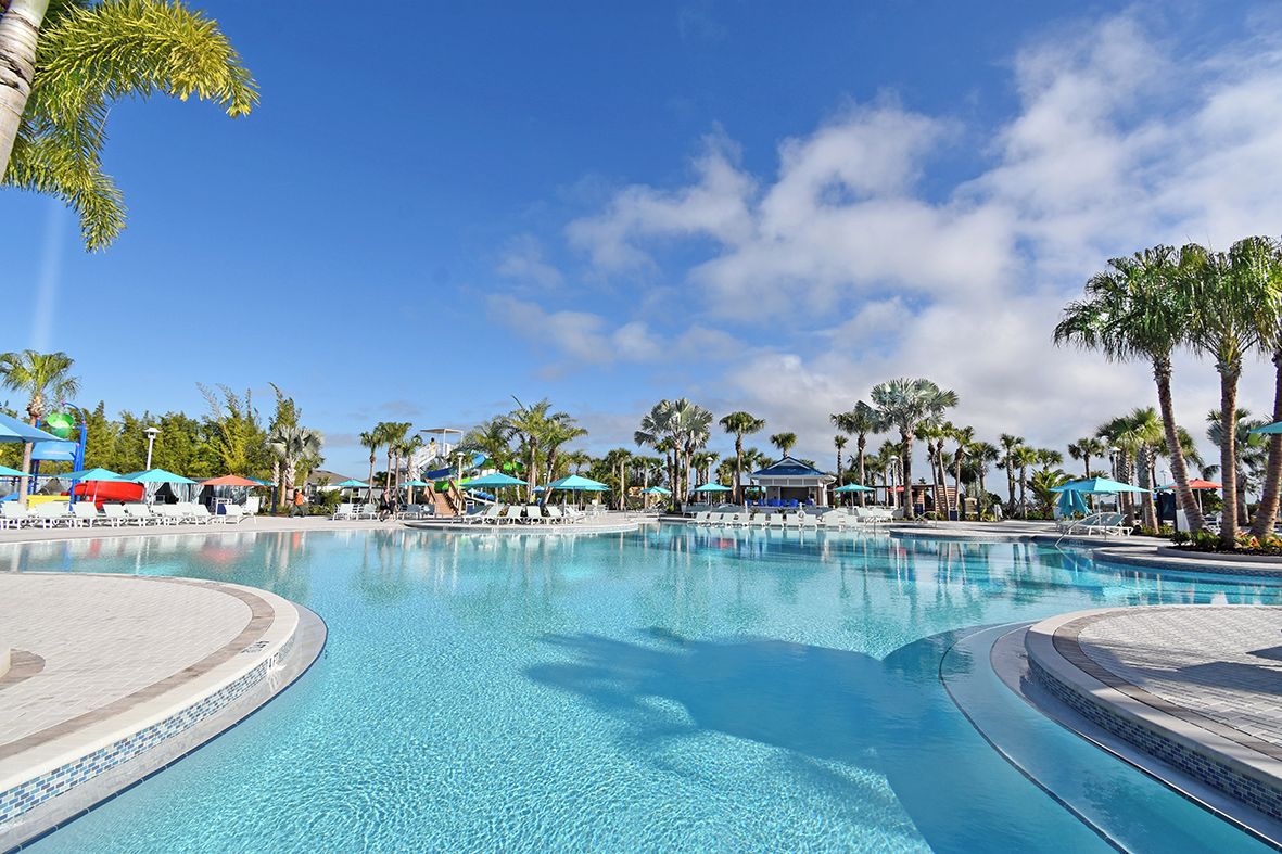 A large outdoor swimming pool with lounge chairs and umbrellas, surrounded by palm trees and clear skies.