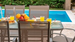 Ontbijt met fruit, sap en ontbijtgranen bij een zwembad, met stoelen en een tafel.