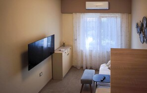 Una habitación compacta con una televisión de pantalla plana montada en la pared, un escritorio de madera pequeño, un banco acolchado y una ventana con cortinas translúcidas.