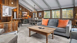 Een ruime woonkamer met houten plafond, een grijze bank met oranje kussens, een houten salontafel en uitzicht op bomen door grote ramen.