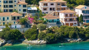 A coastal residential area with multiple two-story buildings, some with balconies, and a clear view of the sea with paddleboarders. - Beachfront Aetheria House, Wonderful Sea Views, Furnished Terrace, Fiskardo (Kefalonia)