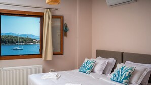 A bedroom with a large window overlooking a body of water, a bed with white bedding and blue pillows, a radiator, and a wall-mounted air conditioner. - Beachfront Aetheria House, Wonderful Sea Views, Furnished Terrace, Fiskardo (Kefalonia)