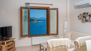 A room with a large window offering a view of the sea and a distant island. There is a television on a wooden stand, a white radiator, and a white sofa with a white knitted throw. - Beachfront Aetheria House, Wonderful Sea Views, Furnished Terrace, Fiskardo (Kefalonia)