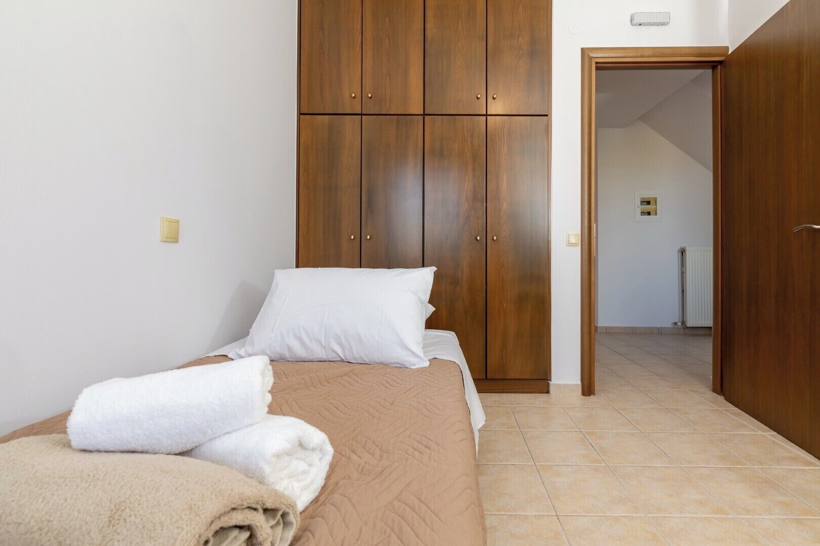 A bedroom with a bed, white pillows, and a brown duvet. There is a wooden wardrobe with two doors and a door leading to another room.