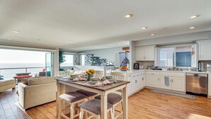 Une cuisine moderne avec des meubles blancs, une table à manger en bois et des tabourets de bar, et une vue sur la mer par de grandes fenêtres.