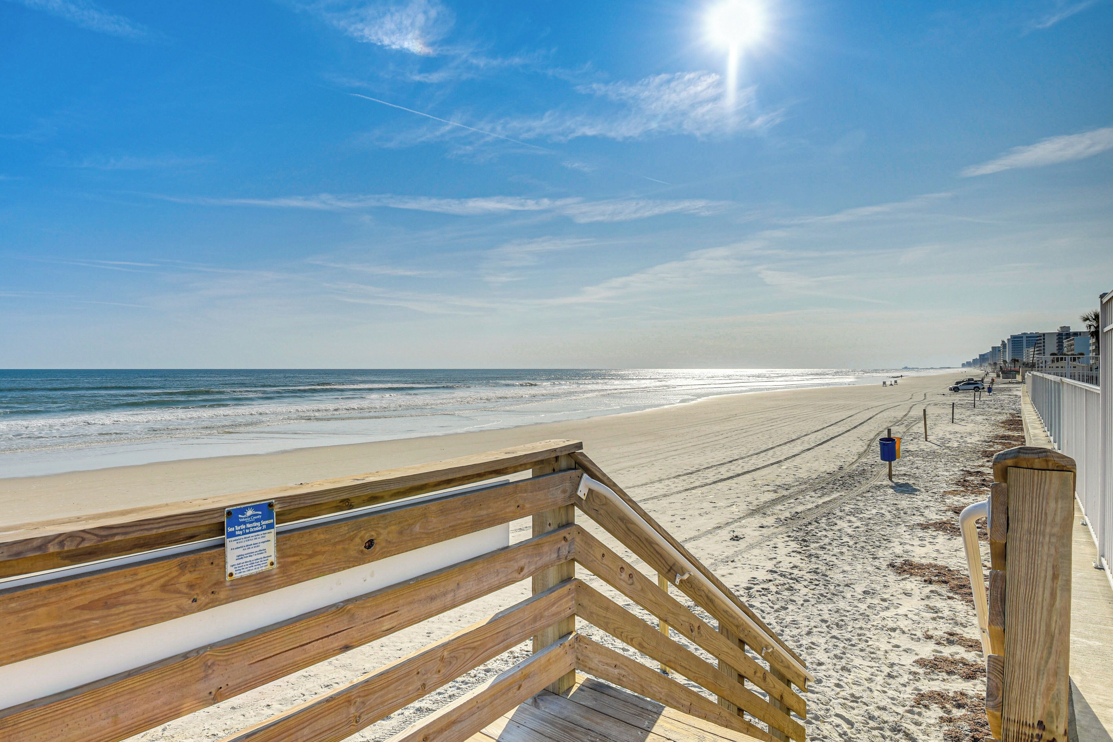 A wooden boardwalk leading to a sandy beach with a clear sky and ocean in the background.