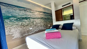 Et soveværelse med et stort mural af et strandmotiv, en seng med hvide sengetøj og et natbord med en lampe.