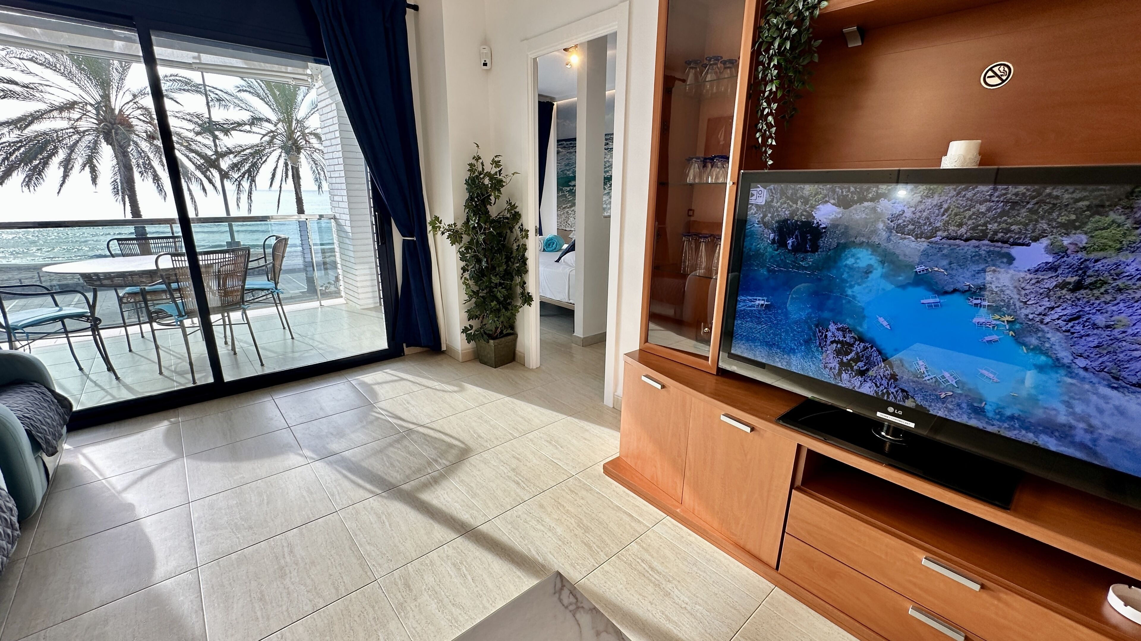 A living room with a large TV displaying a tropical beach scene, a balcony with a view of palm trees and the sea, and a wooden cabinet with drawers.