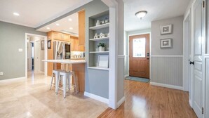 A modern kitchen with white cabinets, a dining area with stools, and a hallway with wooden flooring and a door. - Marbella Lane - Charming 4BR Bungalo for Relaxing Retreat (Fullerton)