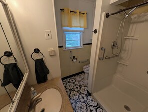 Een badkamer met een wastafel, toilet en douche. Er is een raam met een geel gordijn, en de vloer heeft een betegeld patroon.