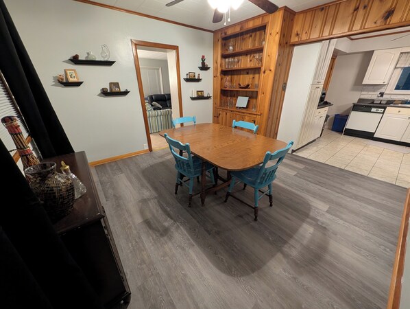 Een eethoek met een houten tafel en blauwe stoelen, een keuken met witte kasten en een houten boekenkast.