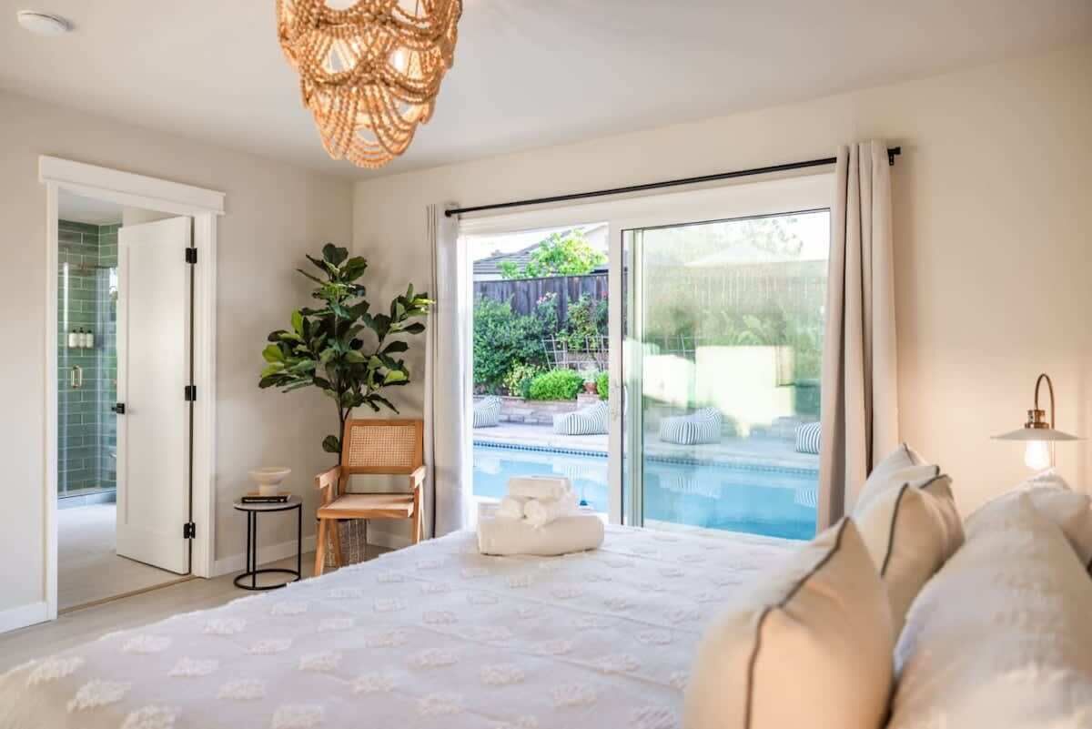 A bedroom with a large bed, a potted plant, a small table, and a view of an outdoor swimming pool.