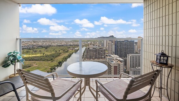 A balcony with a table and chairs, a potted plant, and a view of a cityscape and river. - High-Floor Waikiki Apt w/ Views & Pool (Honolulu)