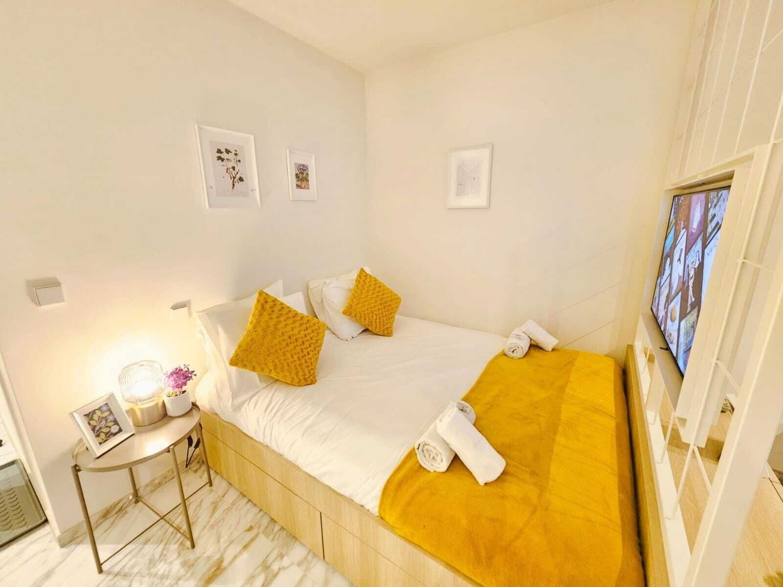 Una habitación de hotel ordenada con una cama, una mesita de noche con lámpara y un televisor instalado en la pared.