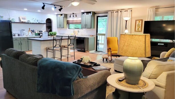 Una sala moderna con sofá, una mesa de centro y televisor. Se observa una zona de cocina con mesa y sillas.